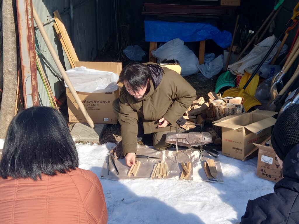 ソロキャンプ体験の活動の様子の写真の2枚目。講師の小山さんが雪の積もった地面の上にしゃがみ、敷物の上に焚き木を並べて説明している場面。